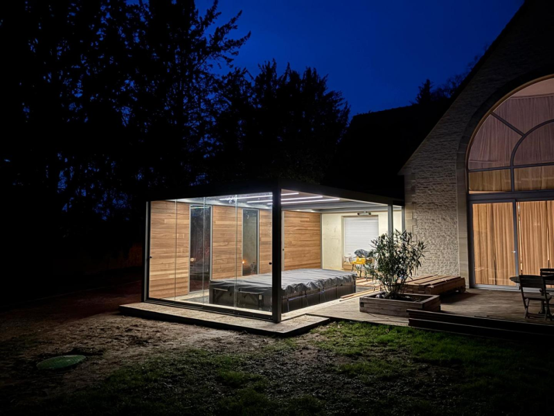 Pergola vitrée anthracite éclairée la nuit, spa couvert et maison en pierre