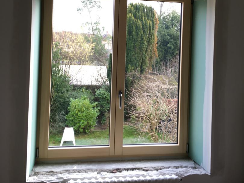 Fenêtre bois clair à deux vantaux en cours de pose, radiateur blanc et jardin