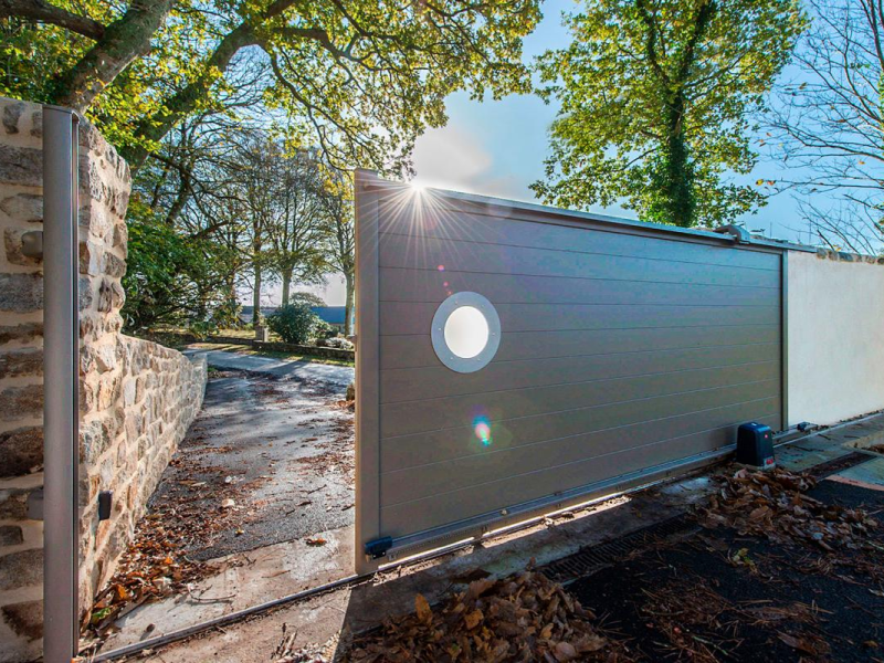Portail coulissant gris-taupe à lames horizontales et hublot rond, mur pierre