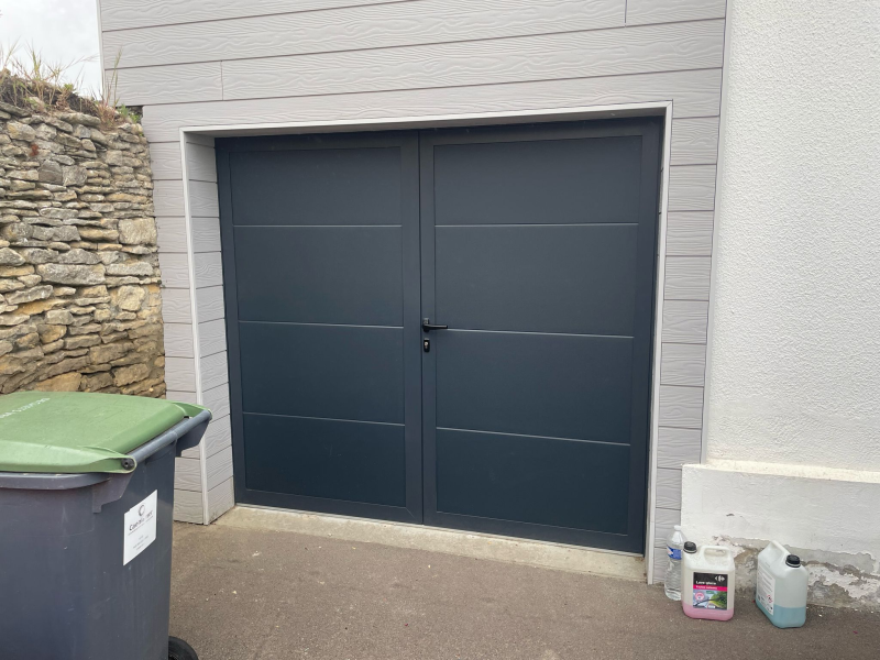 Porte de garage battante anthracite à deux vantaux, bardage gris et mur pierre