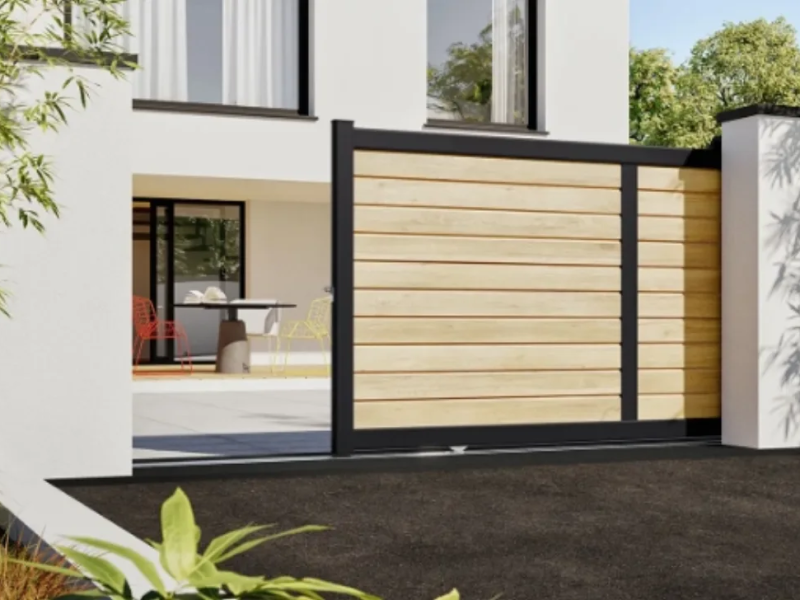 Portail coulissant à lames aspect bois clair et cadre noir devant maison blanche