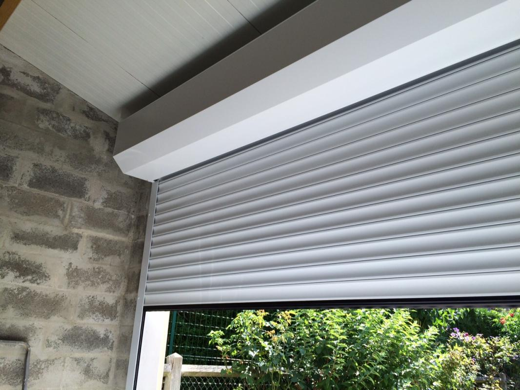 Volet roulant blanc partiellement baissé sur mur en parpaings, vue sur jardin