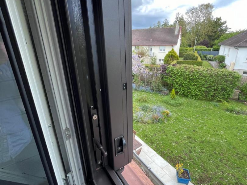 Volet pliant aluminium anthracite sur fenêtre ouverte, vue jardin et maison