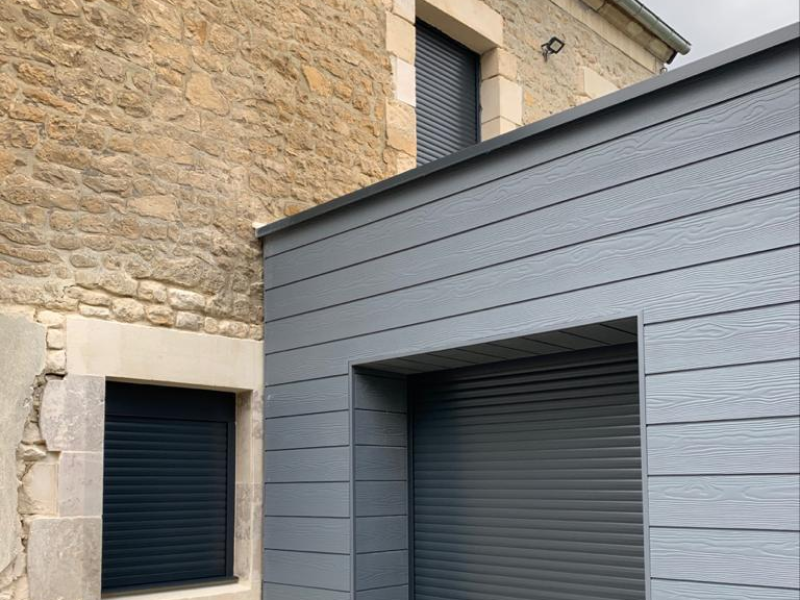 Extension à bardage anthracite et volets roulants fermés sur mur en pierres