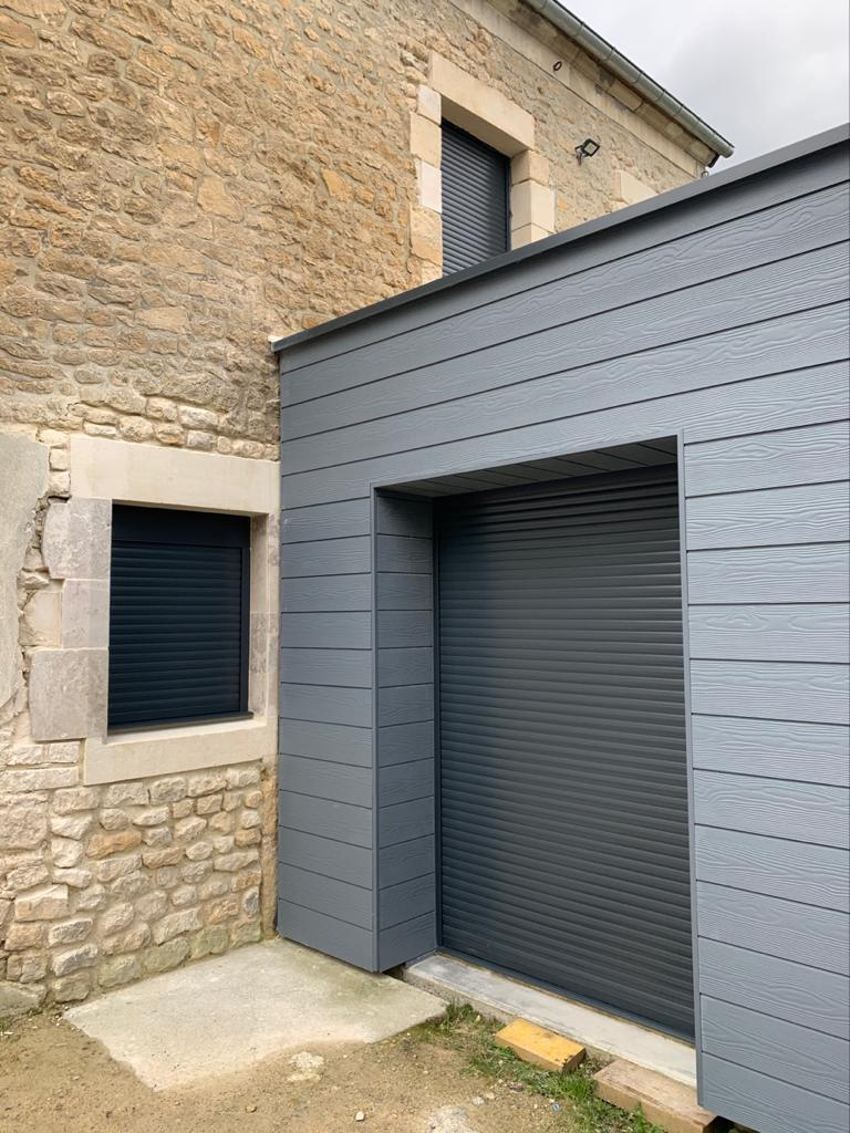 Extension à bardage anthracite et volets roulants fermés sur mur en pierres